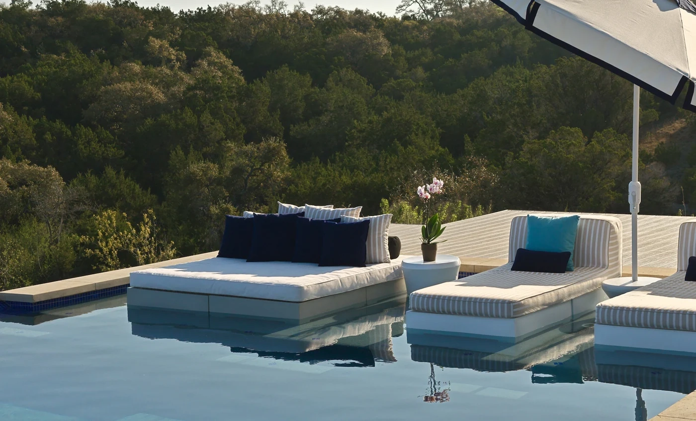 Luxurious poolside setting with plush loungers and cushions by an infinity pool, surrounded by lush greenery. A white parasol and potted orchid add elegance.