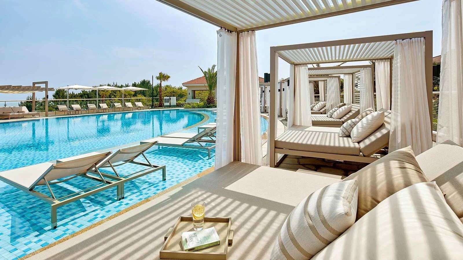 Luxurious poolside cabanas with white drapes and plush loungers overlook a sparkling blue pool. Relaxing atmosphere, with a refreshing drink on a side table.
