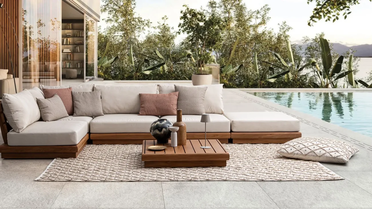 Modern outdoor patio with a beige sectional sofa and assorted pillows, set beside a pool. Wooden coffee table with decor atop a woven rug, lush greenery in the background.