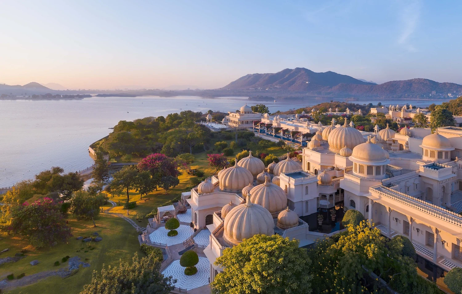 Aerial view of a grand palace complex with ornate domes and lush gardens beside a serene lake. Mountains and a sunrise glow softly in the background.