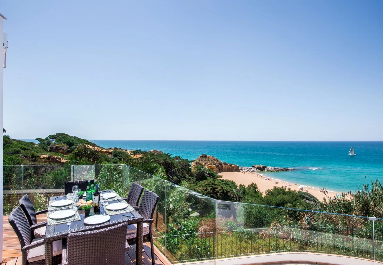 A scenic coastal view from a balcony with a set dining table. Overlooks a sandy beach, turquoise ocean, and a distant sailboat under a clear sky.