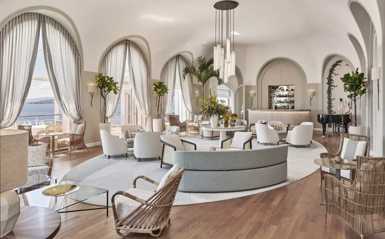 Luxurious lounge at the hotel du cap eden roc with arched windows overlooking the ocean. Elegant round sofas, wicker chairs, and a grand piano create a serene, sophisticated ambiance.