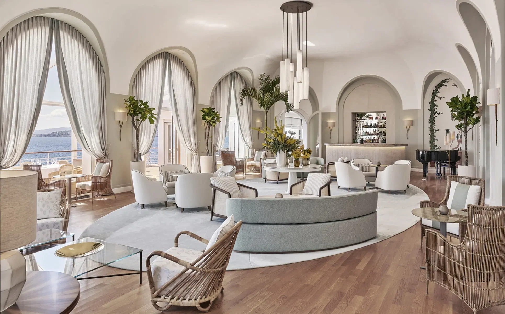 Luxurious lounge at the hotel du cap eden roc with arched windows overlooking the ocean. Elegant round sofas, wicker chairs, and a grand piano create a serene, sophisticated ambiance.