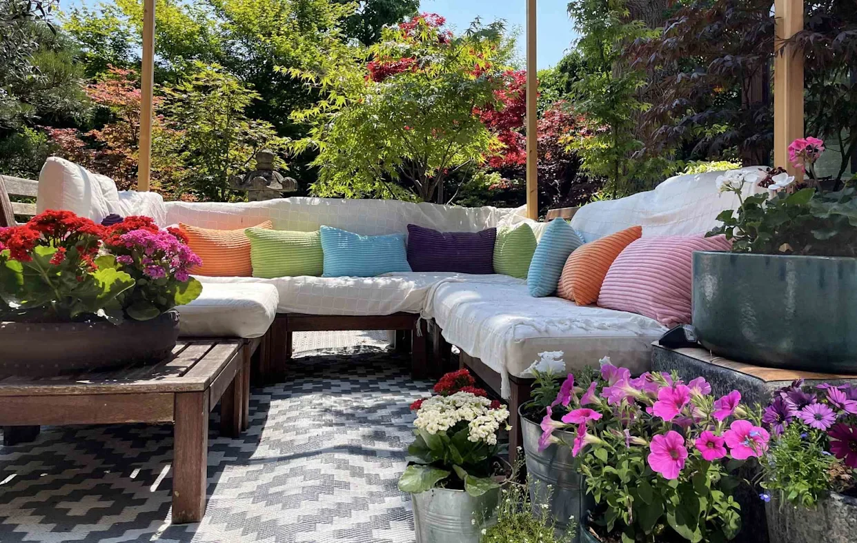 Outdoor patio with a sectional sofa covered in white fabric, adorned with colorful throw pillows. Surrounding flowers in bloom create a vibrant, serene ambiance.