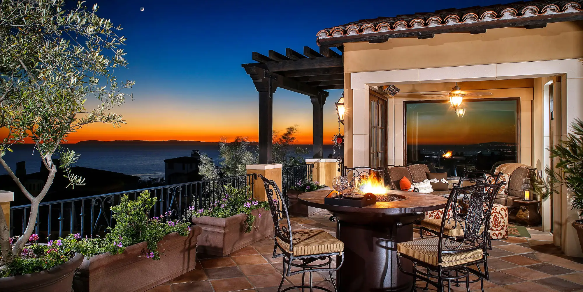 Cozy terrace at sunset overlooking the ocean. Features a fire pit table and ornate chairs under a wooden pergola. Warm, inviting ambiance, common in luxury rooftops.