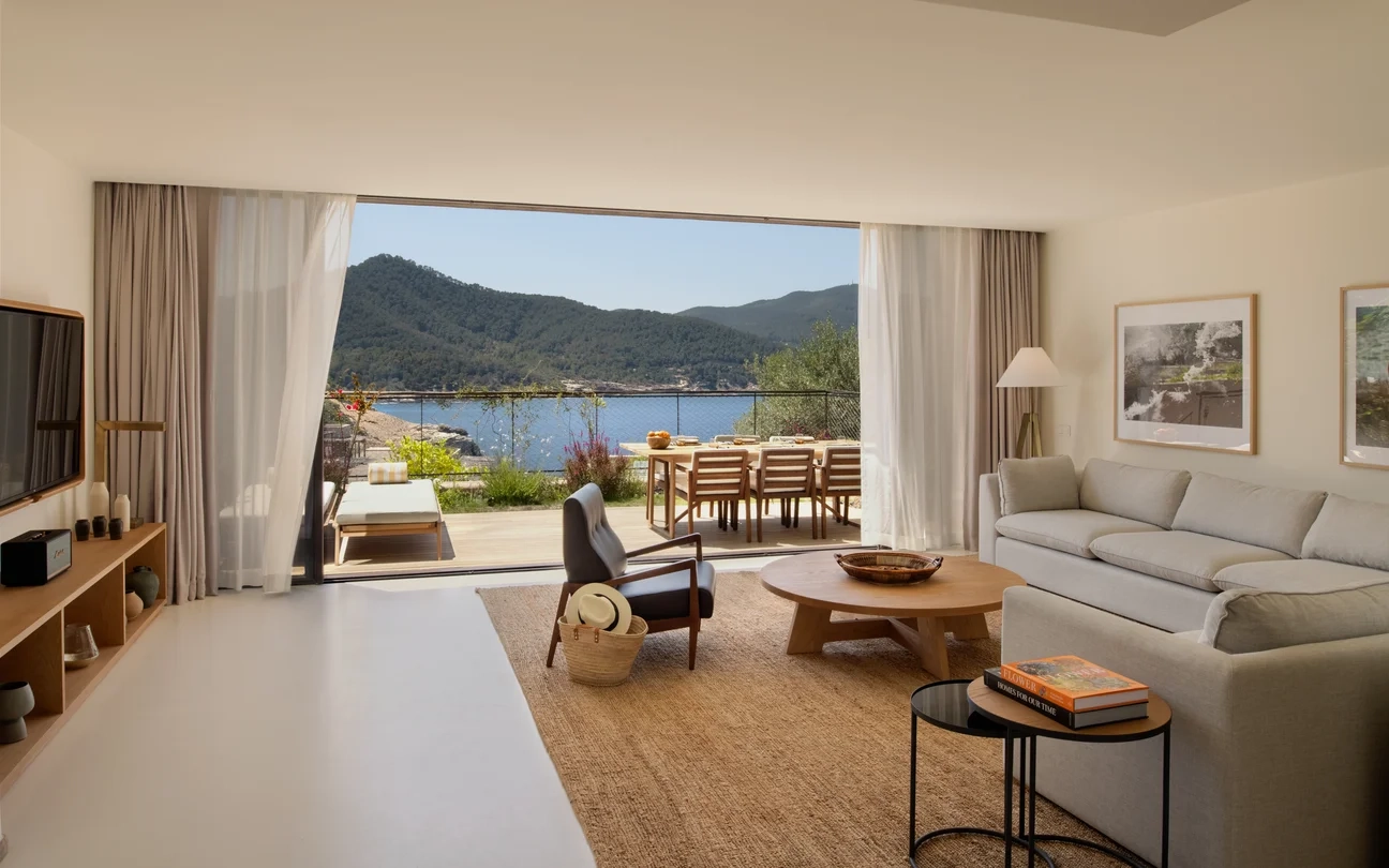 Spacious living room with a large glass door opening to a patio at the Siz Senses Ibiza. Soft gray sofa, wooden furniture, and coastal view create a serene, inviting atmosphere.