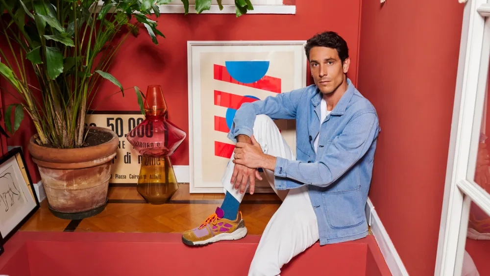 Interior designer Francesco Meda in a light blue jacket and white pants sits on a red step indoors, surrounded by modern art, vibrant plants, and colorful decor. 
