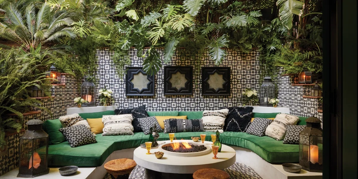 A cozy outdoor lounge with lush greenery, a U-shaped green sofa, patterned cushions, a round fire pit, lanterns, and patterned walls, creating a serene ambiance.
