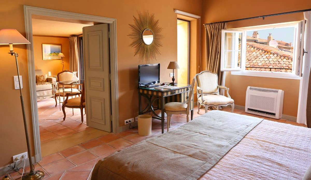 A warm, cozy suite at the Hotel La Ponche in Saint-Tropez with beige walls features a bed, a desk with a TV, and an open window showing a tiled roof. 