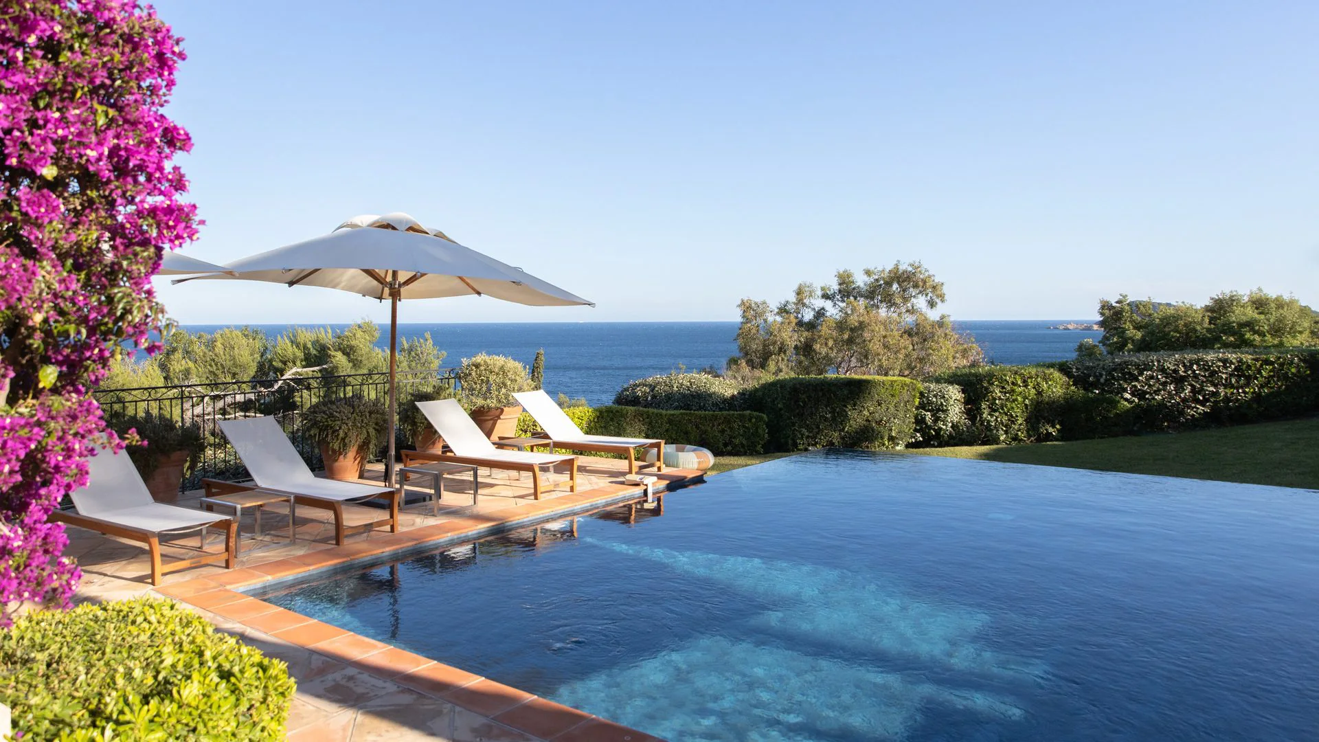 Infinity pool at the Reserve Ramatuelle with wooden lounge chairs under a white umbrella, overlooking a serene ocean view. Bougainvillea blooms and lush greenery add vibrant colors.