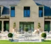 Elegant stone house with large windows and a spacious patio featuring white furniture and manicured topiary in planters, exuding a serene, upscale vibe.