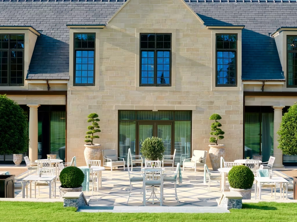 Elegant stone house with large windows and a spacious patio featuring white furniture and manicured topiary in planters, exuding a serene, upscale vibe.
