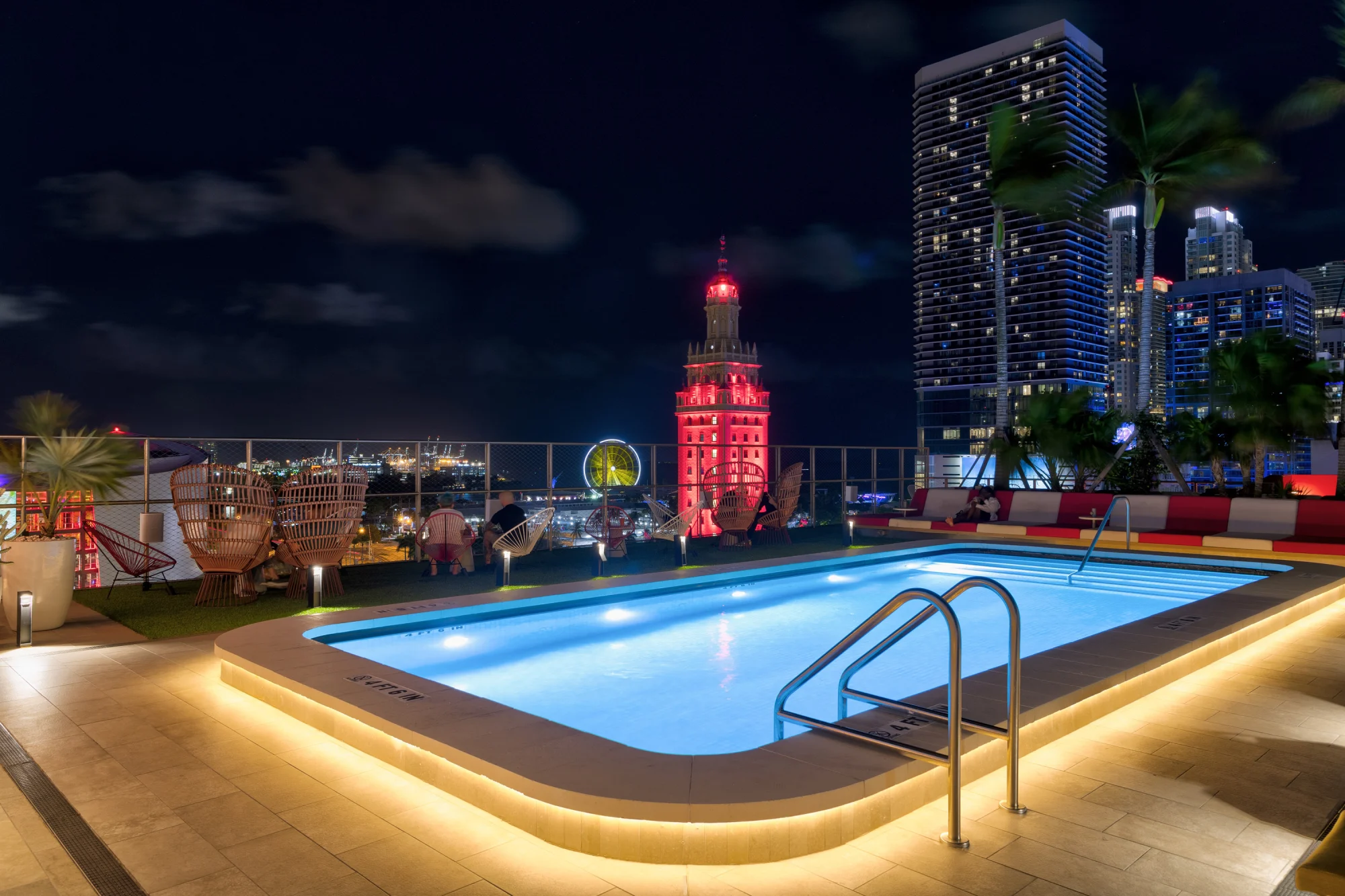 Rooftop pool at night with city skyline; features glowing blue water, loungers, red-lit historic building, and modern high-rises under a cloudy sky, highlighting the luxurious pool decks in Miami.