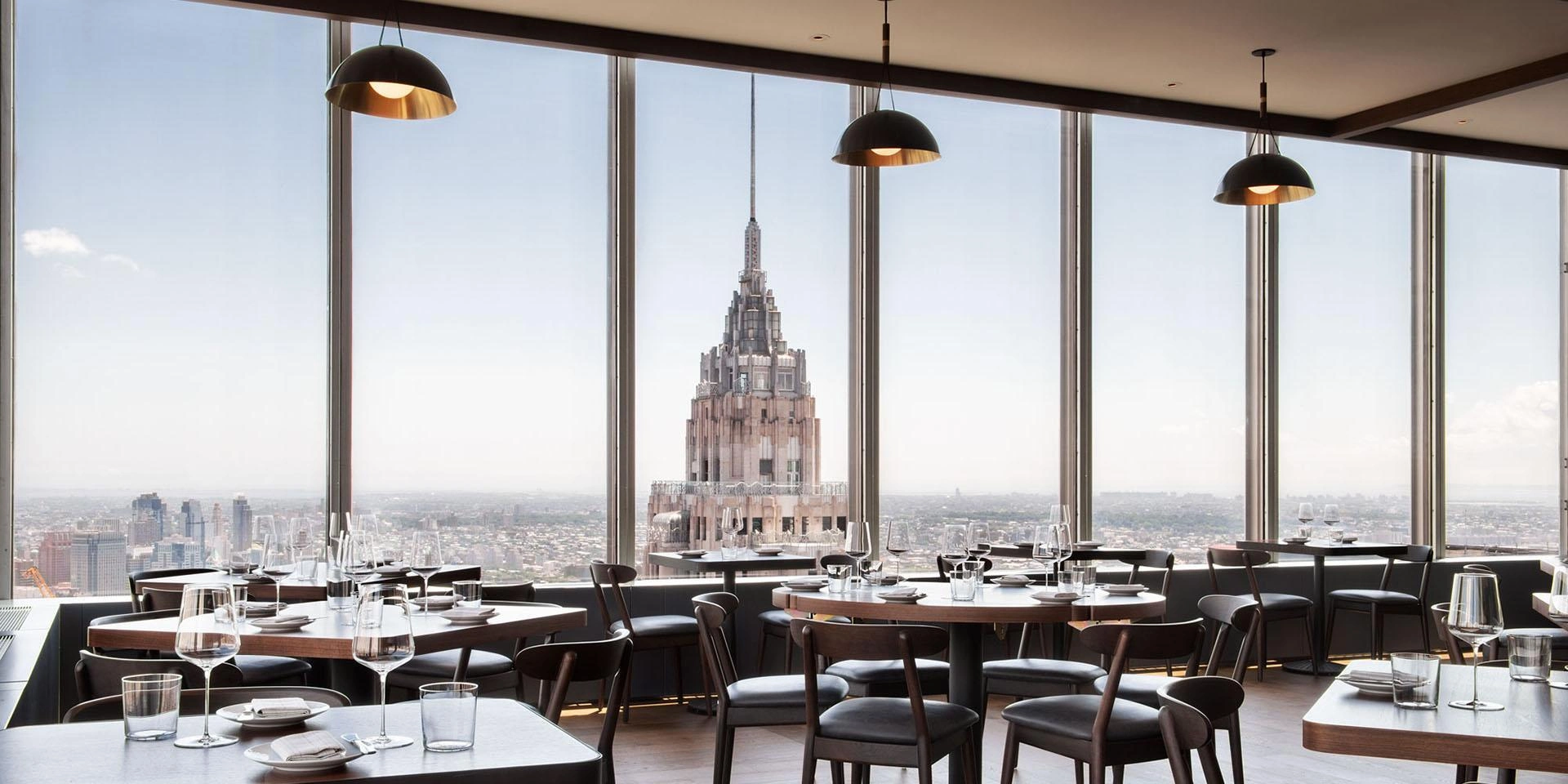 Modern rooftop restaurant interior in New York with large windows offering a panoramic city view. Elegant tables and chairs create a sophisticated, serene ambiance.
