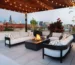 Rooftop patio with modern sofas and armchairs around a lit fire pit. Overhead string lights, potted plants, and a city skyline create a cozy atmosphere.