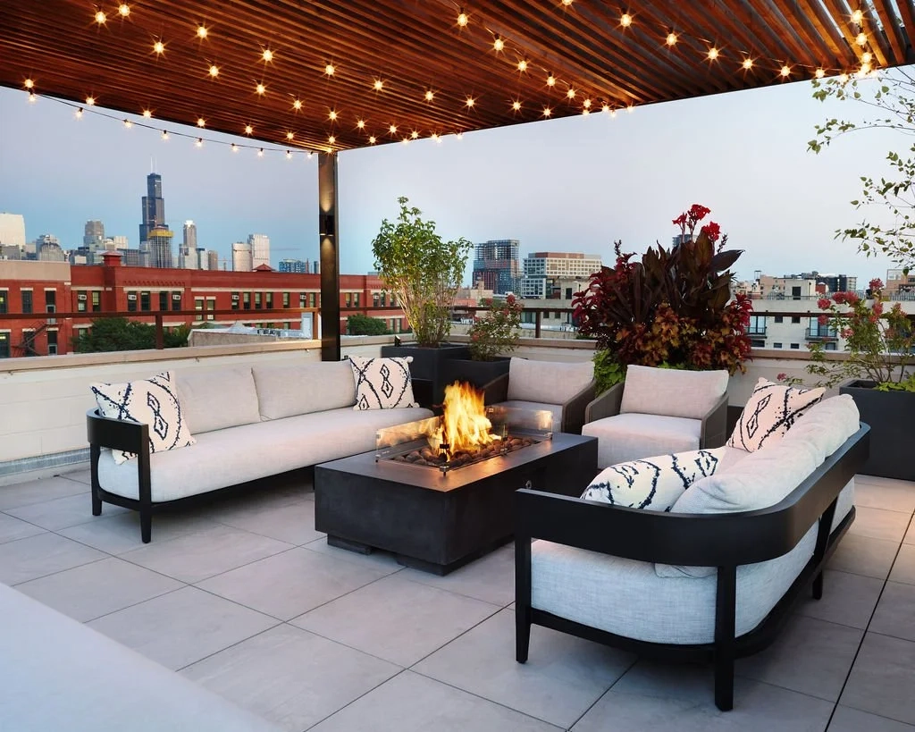 Rooftop patio with modern sofas and armchairs around a lit fire pit. Overhead string lights, potted plants, and a city skyline create a cozy atmosphere.