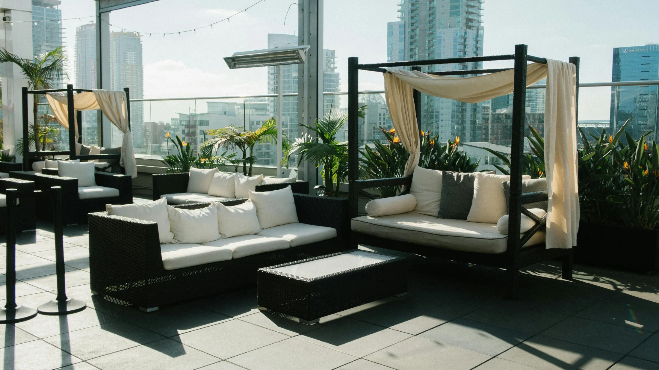 Rooftop lounge with cozy wicker sofas, cream cushions, and canopy drapes. Surrounded by potted plants and city skyscrapers under clear skies. Calm ambiance.