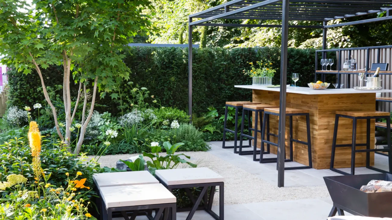 A cozy garden patio with an outdoor home bar under a pergola, surrounded by lush greenery and colorful flowers, creating a serene atmosphere.