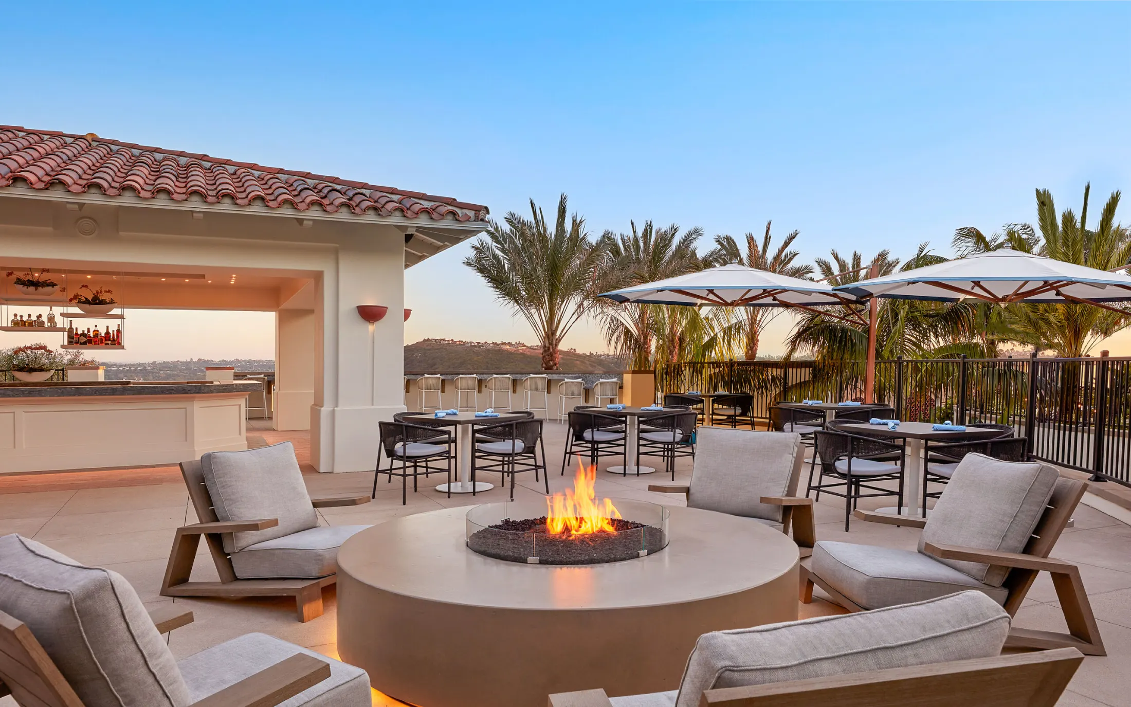 Rooftop patio at sunset with a cozy fire pit, surrounded by cushioned chairs. Palm trees and open umbrellas add a tropical, relaxing ambiance.