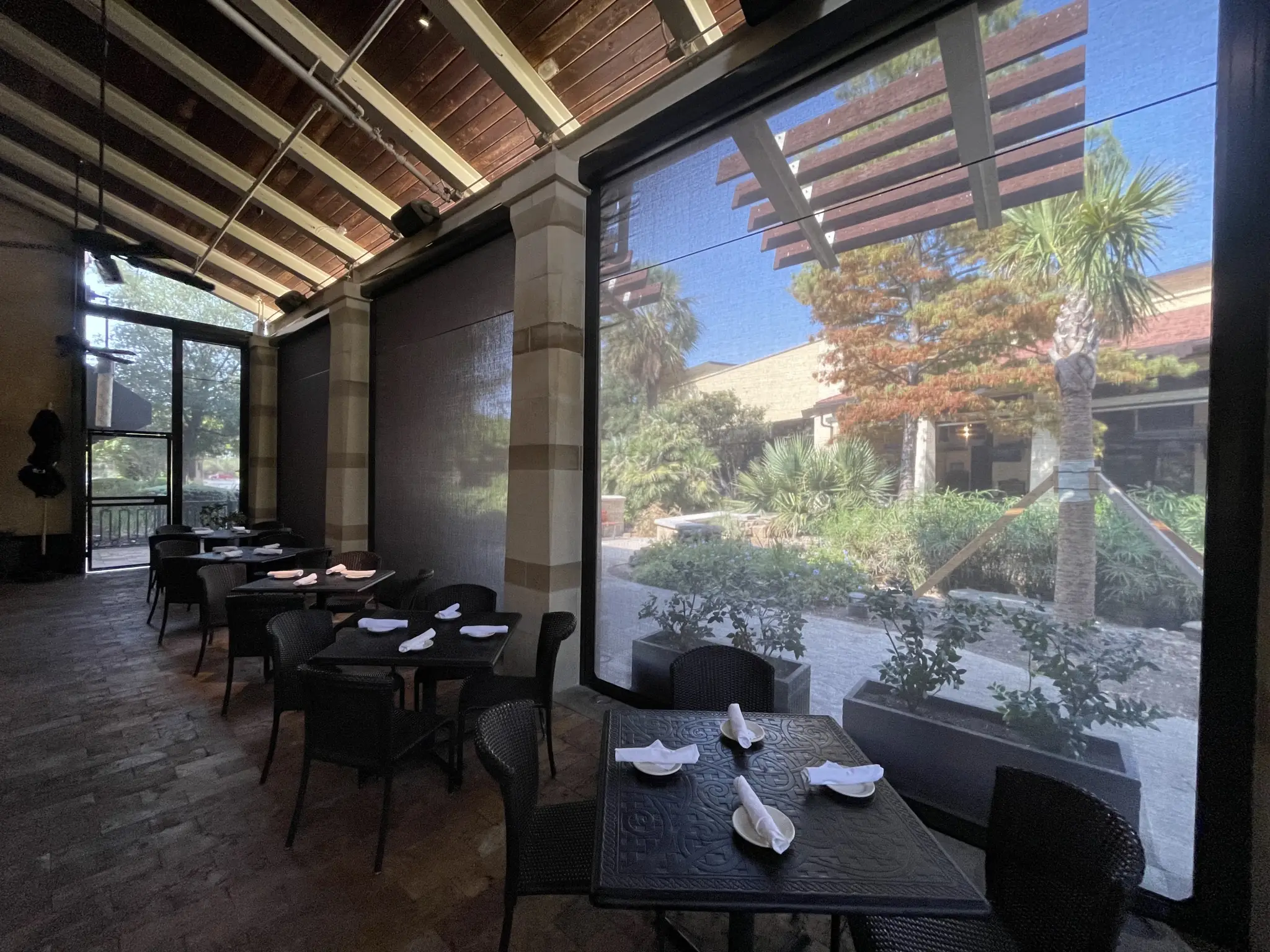 Cozy restaurant patio with dark wicker chairs and neatly set tables, overlooks a sunlit garden with lush greenery and tall palm trees outside.