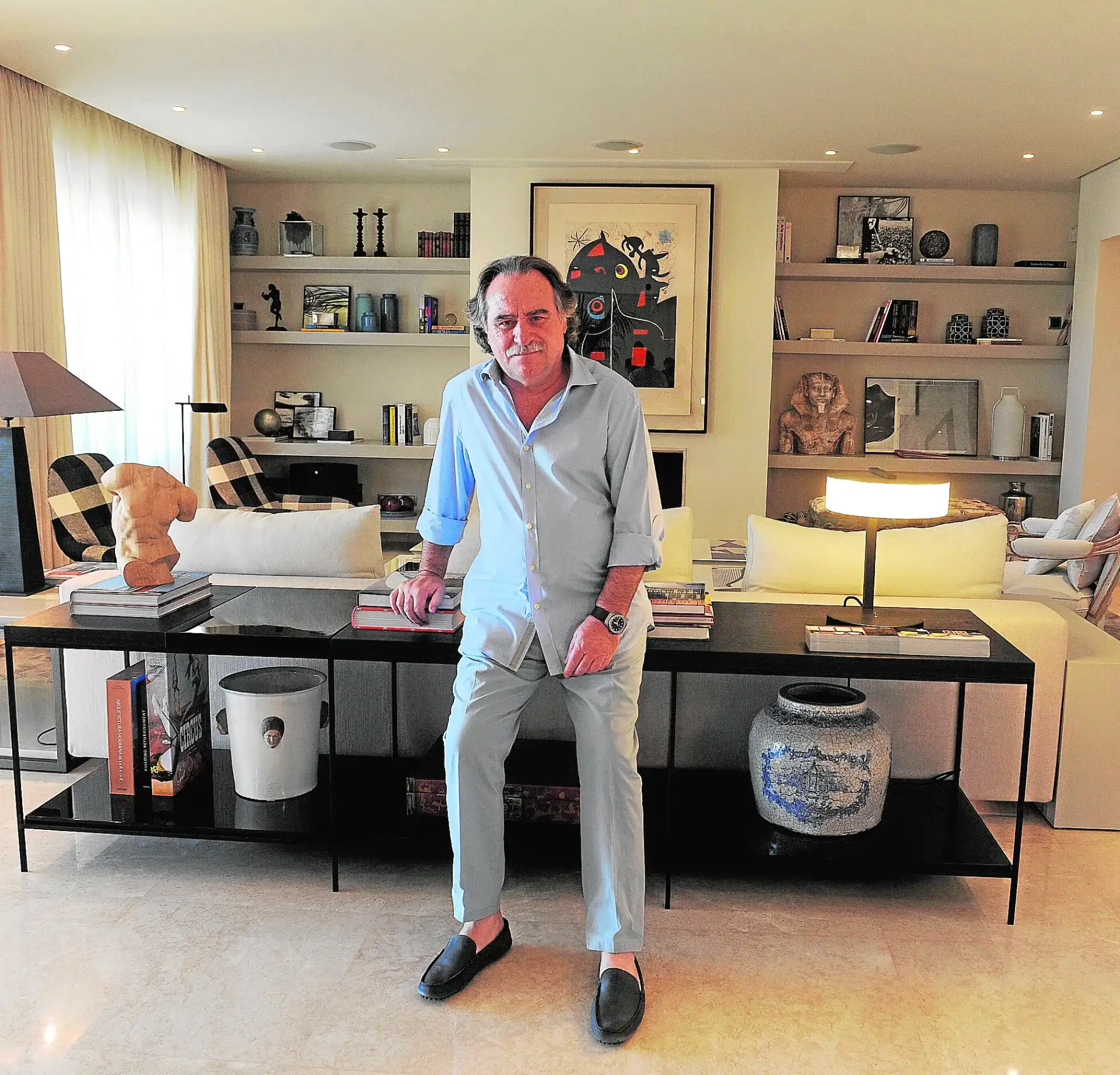 Interior designer Pedro Peña in a light blue shirt and pants leans on a console table in a stylish living room. Books, art, and a sculpture create an elegant ambiance.