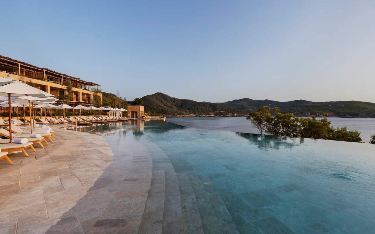 Luxurious infinity pool overlooking a tranquil bay, surrounded by sun loungers and umbrellas on a stone deck. Hills and serene water create a calm atmosphere.