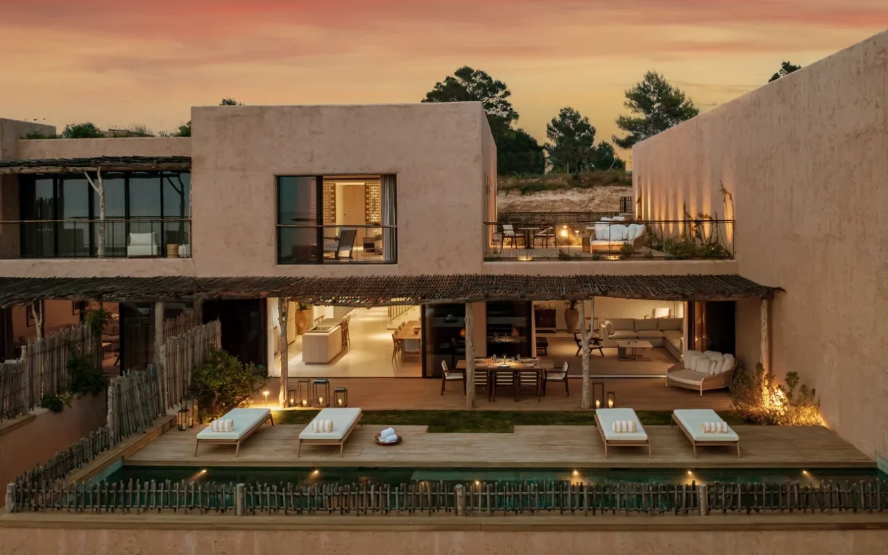 Siz Senses Ibiza, Spain, at sunset with modern architecture, featuring large windows, an outdoor dining area, poolside loungers, and warm ambient lighting.