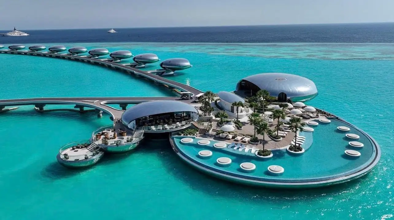 Futuristic Shebara Resort with pod-like structures connected by walkways over turquoise water. Lush greenery and lounge chairs on deck; calm, tropical vibes.