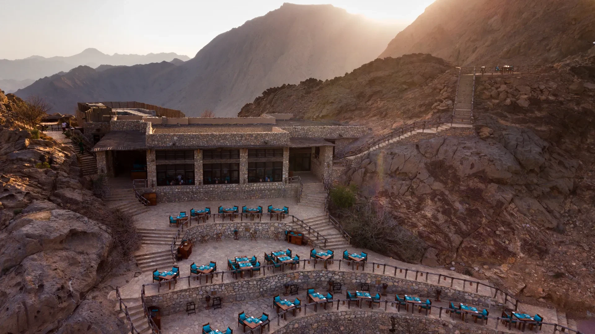 A stone terraced restaurant at the Six Senses Zighy Bay set on a rocky hillside during sunset. Tables with blue tablecloths overlook mountain views, creating a serene atmosphere.