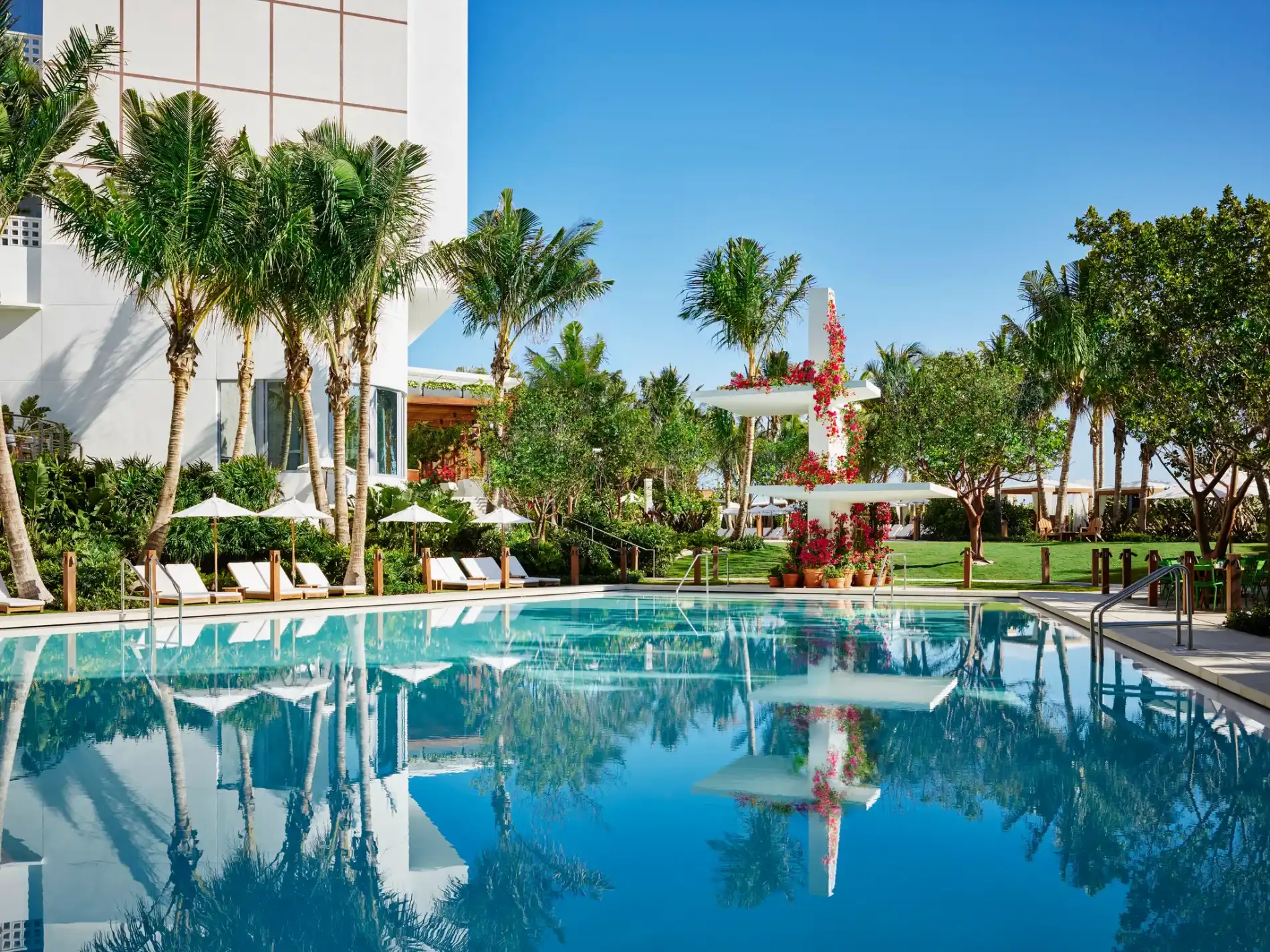 A luxurious pool reflecting clear blue skies, surrounded by palm trees, lounge chairs, and lush greenery. A serene and inviting tropical oasis at the Miami Beach Edition.