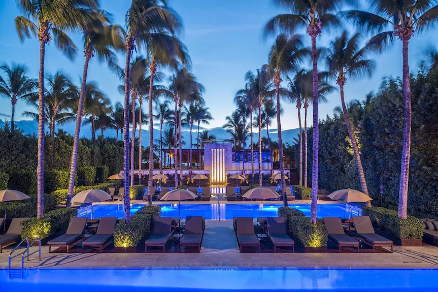 Luxurious resort pool at The Setai in Miami at twilight, surrounded by tall palm trees, lounge chairs, and umbrellas. Ambient blue lighting creates a serene, inviting atmosphere.