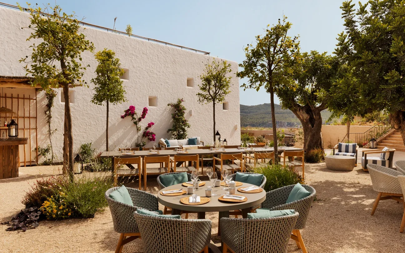 Outdoor dining area at The Orchard, Six Senses Ibiza, with rustic charm, featuring round tables set for a meal. Surrounded by trees and a white stucco wall, evoking a relaxed, elegant vibe.