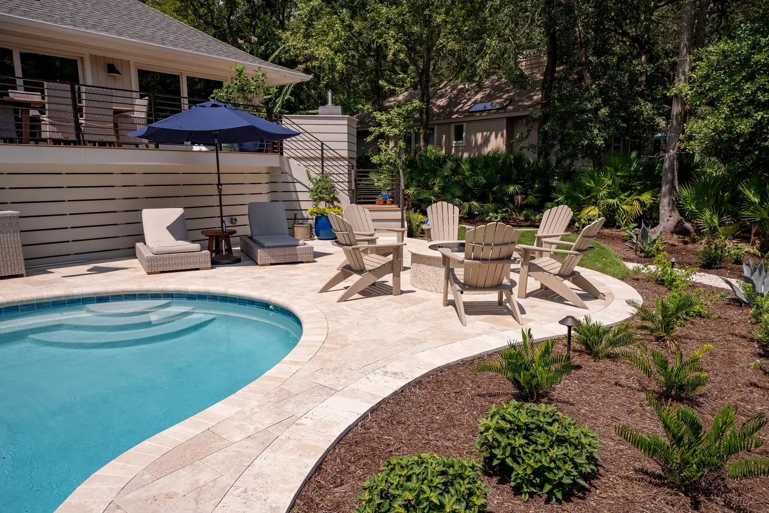 Luxurious travertine stone patio with a blue pool, surrounded by lounge chairs, fire pit with Adirondack chairs, lush greenery, and shaded by trees, creating a serene vibe.